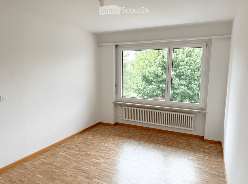 Empty room with wooden flooring, white walls, and a large window with a radiator below it.