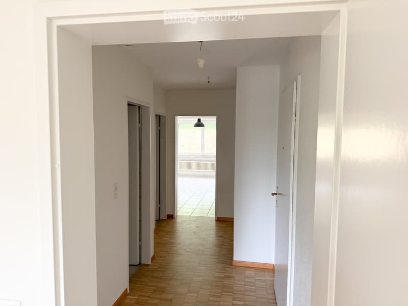 Bright hallway with wooden flooring, white walls, and multiple doors leading to other rooms. Natural light streams in from a window at the end of the corridor.