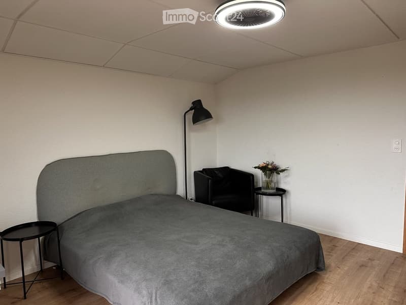 A modern bedroom with a grey bed, a black armchair, a small black side table with a vase of flowers, and a floor lamp. The room features light wooden flooring and white walls.