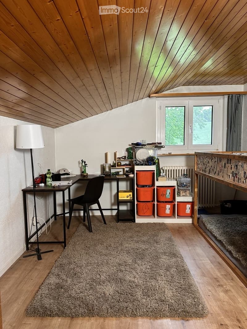 Cozy attic room with wooden slanted ceilings, a desk with a chair, a floor lamp, a shelf with storage bins, and a bed.