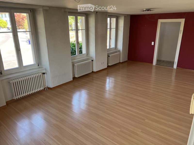 Empty room with wooden flooring, white radiators beneath windows, and a red accent wall. No furniture or equipment present.