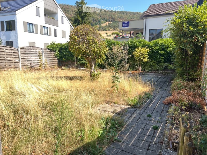 Garden area with a paved pathway, overgrown grass, and surrounding greenery, with two modern houses in the background.