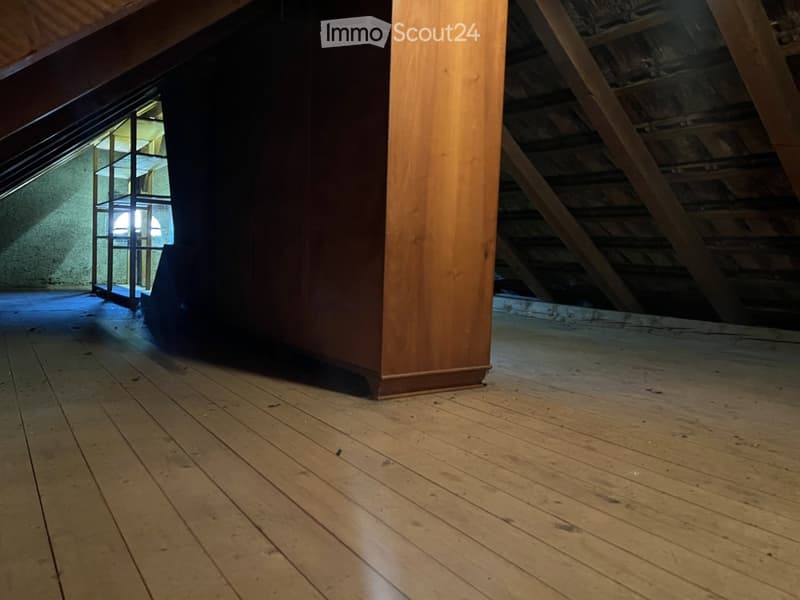 Empty attic space with wooden flooring, exposed wooden beams, and a small window letting in light.