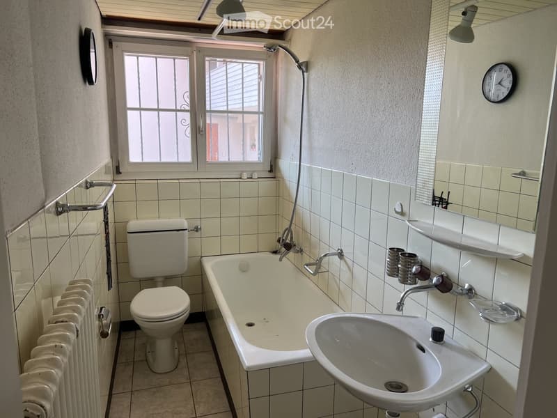 Bathroom with a toilet, bathtub, sink, radiator, mirror, and clock. The walls are tiled, and there is a window with bars.