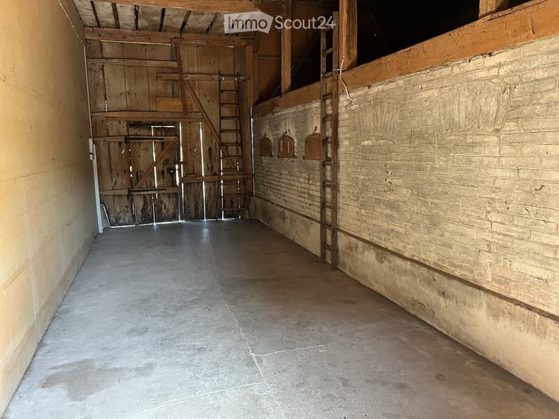 Empty room with exposed wooden beams, brick walls, and a concrete floor. Features a wooden ladder leading to an upper level and a large wooden door.