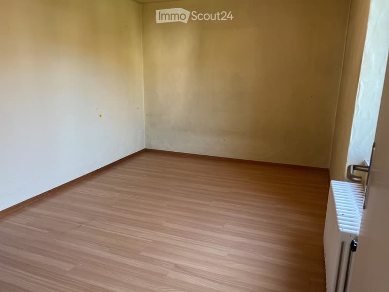 Empty room with wooden flooring and light-colored walls, featuring a radiator and a door frame visible on the right side.