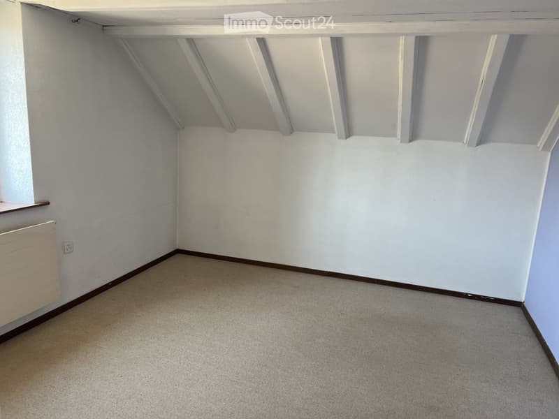 Empty room with white walls, carpeted floor, exposed wooden beams on the ceiling, and a radiator on the left side.