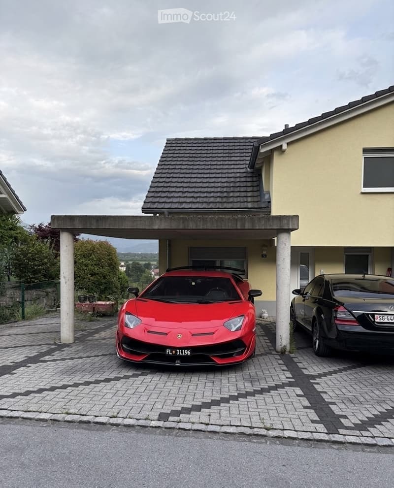 Two-car garage with a red Lamborghini and a black sedan parked inside, attached to a yellow house with a tiled roof.