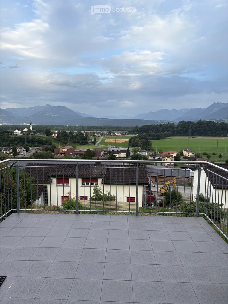 Balcony with tiled floor and metal railing, overlooking a town with houses, fields, and distant mountains.