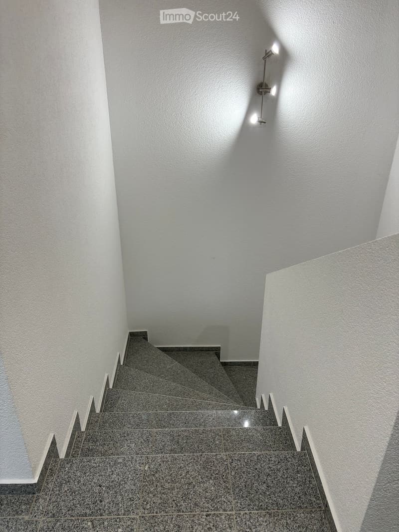 Modern staircase with grey tiled steps and white walls, illuminated by wall-mounted spotlights.