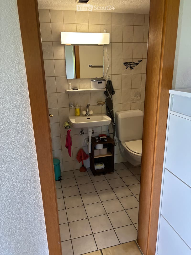 A small bathroom with beige tiles, a white sink, a toilet, a mirror, a small wooden shelf holding toiletries, and a towel hanging on the wall.