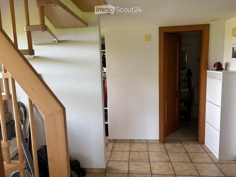 Hallway with tiled floor, wooden staircase, open door leading to bathroom, and a vacuum cleaner placed near the stairs.