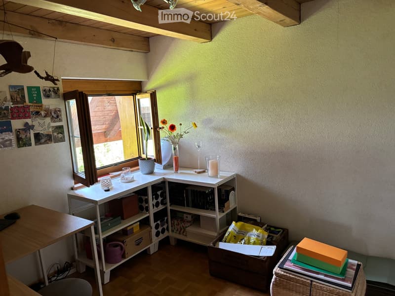 A cozy, well-lit room with wooden flooring, a large window, a small table with a vase of flowers, a shelf with books and storage boxes, and a wicker stool with books and a box on top.