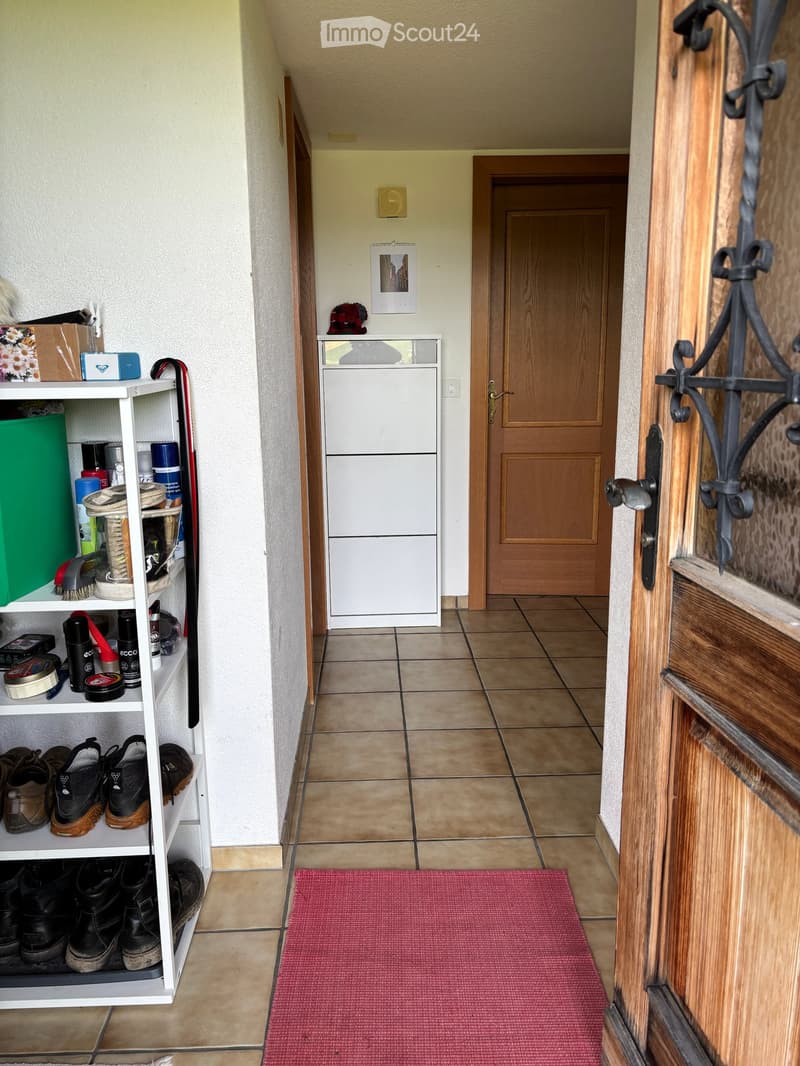 Hallway with tiled floor, wooden door, shoe rack, and small shelf with various items.