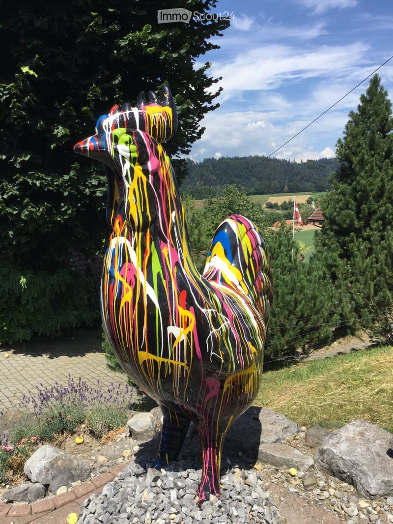 A colorful painted rooster sculpture stands on a bed of rocks in a garden setting, surrounded by greenery and a scenic view of hills and trees.