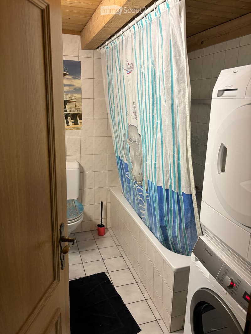 Bathroom with a bathtub, shower curtain, toilet, washing machine, and a black bath mat on the floor.