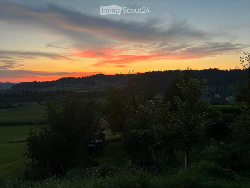 A panoramic view of a rural landscape at sunset, featuring rolling hills, fields, and scattered trees.