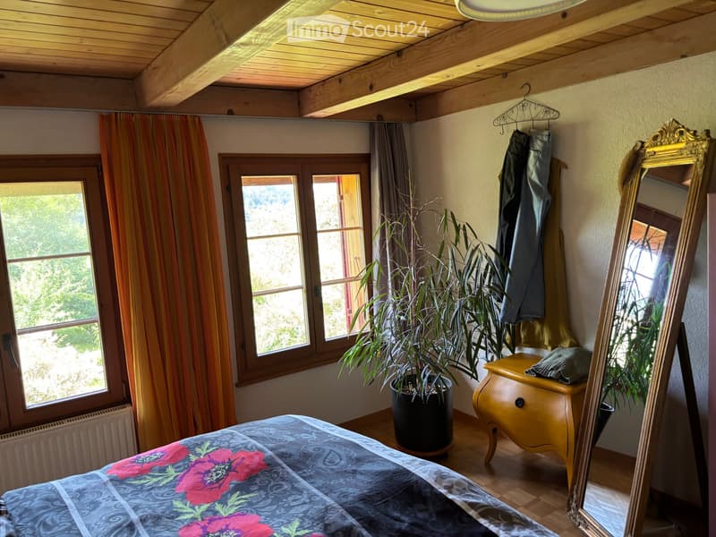 Cozy bedroom with wooden ceiling beams, large windows with striped curtains, a bed with a floral duvet, a potted plant, a nightstand, and a large mirror.