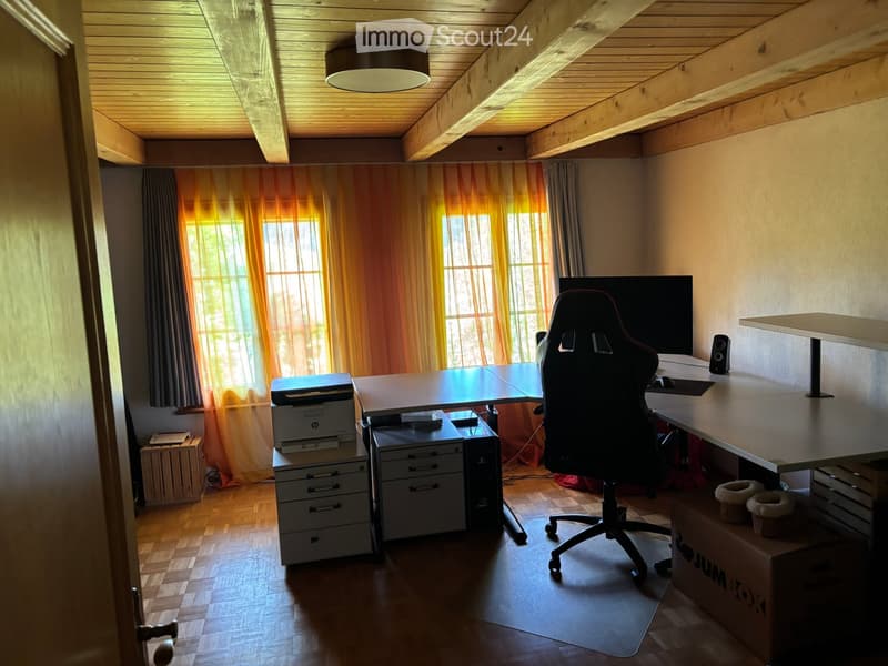 A well-lit room with wooden flooring and a slanted wooden ceiling. It features a large desk with a chair, a printer, a computer tower, a monitor, and a speaker. There are also drawers and a curtain covering the windows.