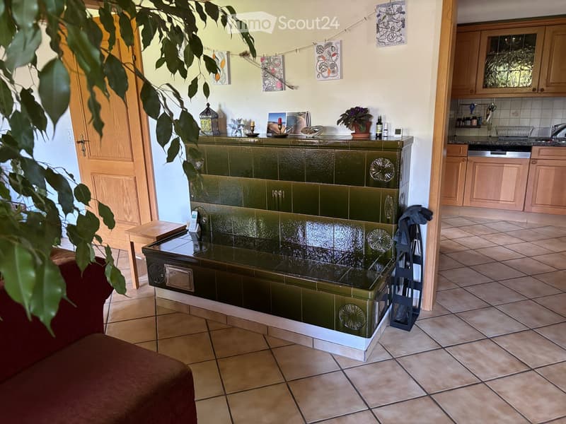 A cozy living room with a green-tiled fireplace, a red sofa, a small wooden table, and a kitchen area with cabinets, a sink, and a dishwasher.