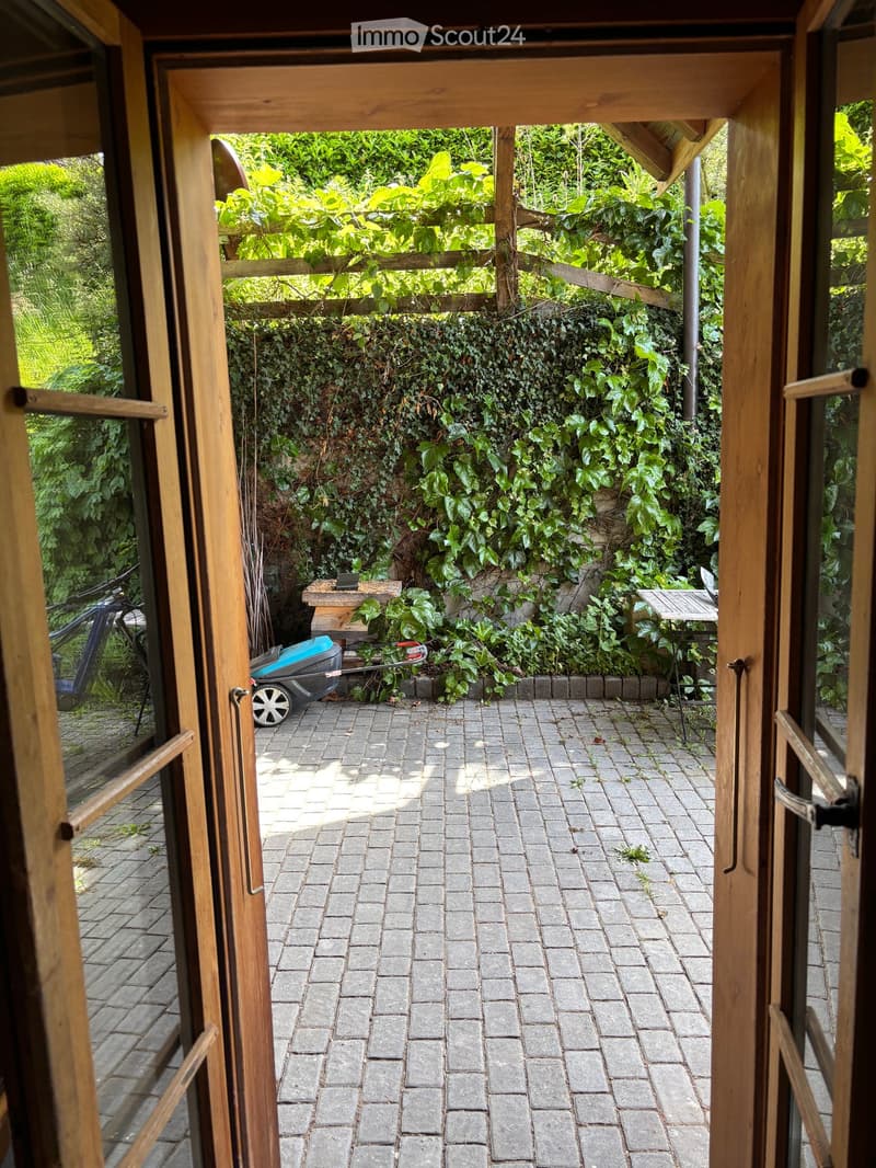 Brick-paved patio area with wooden furniture, greenery, and a small table and chairs.
