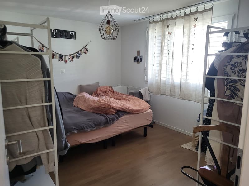 Cozy bedroom with a single bed, wooden floor, white walls, and a hanging pendant light. Features a wardrobe, a chair, and decorative picture frames.