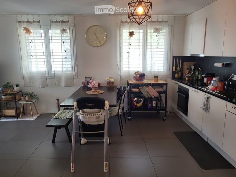 A modern, well-lit kitchen and dining area with a large dining table, chairs, a baby chair, a small bench, and a kitchen cabinet with an oven. The space features a clock on the wall, curtains on the windows, and several potted plants on shelves and tables