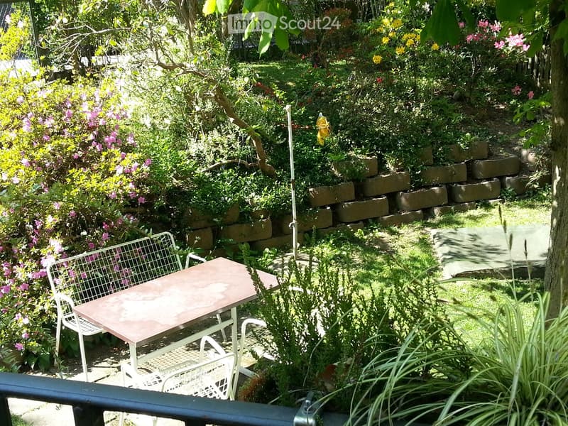A garden with different varieties of flowers, bushes, and grasses. There is a wooden table with white chairs.