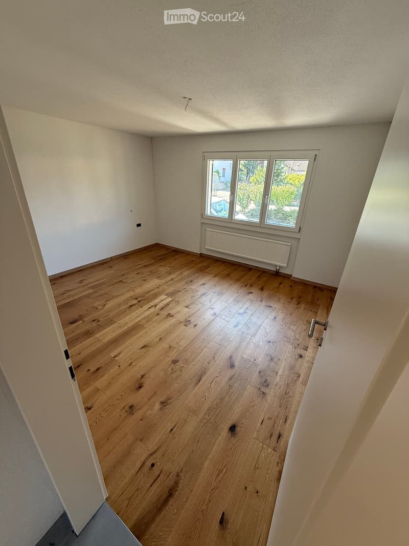 Empty room with wooden flooring, white walls, and a window with three panes. There is a radiator below the window and a door visible on the left side.