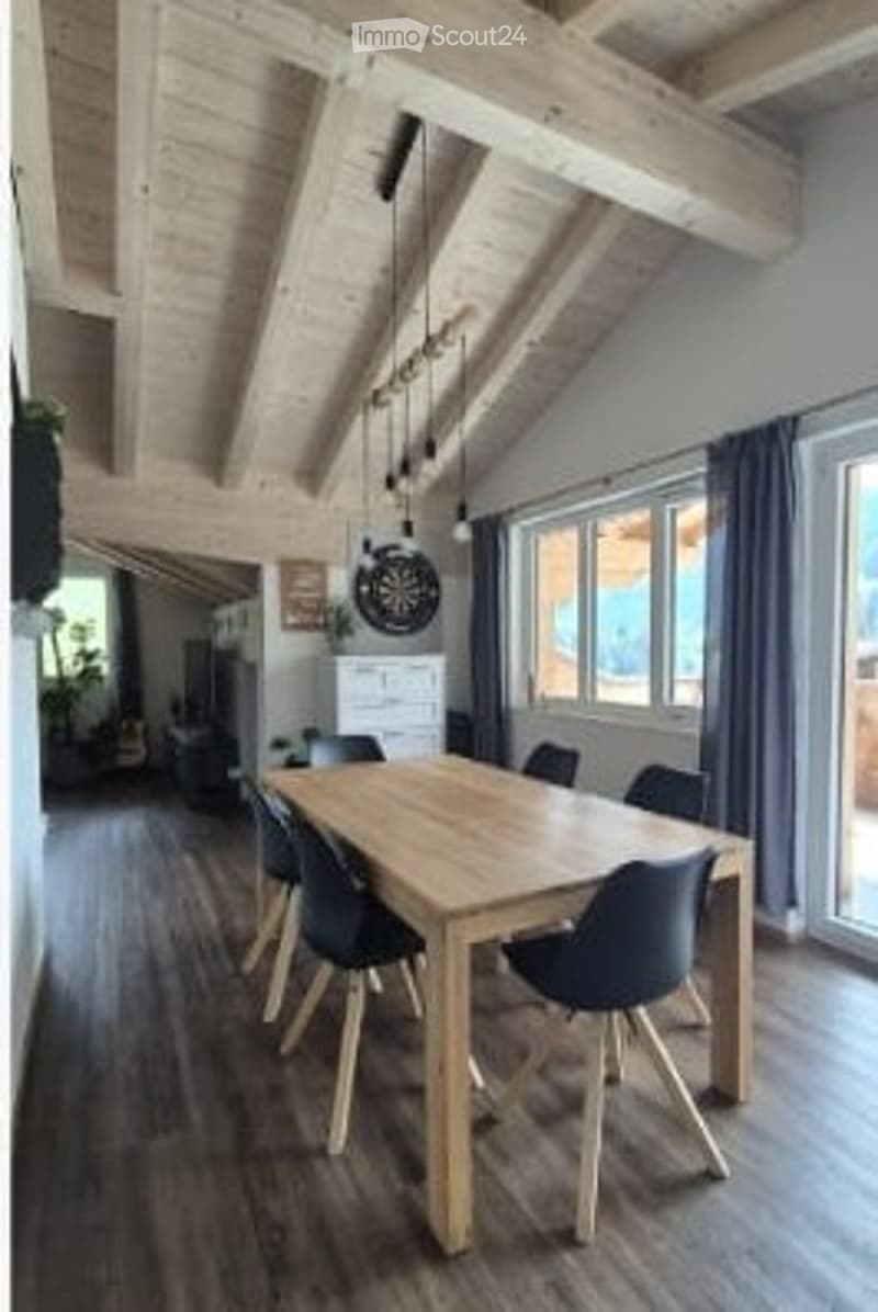 Bright dining room with wooden ceiling beams, large windows, a wooden dining table with black chairs, a white cabinet, and a decorative picture frame on the wall.