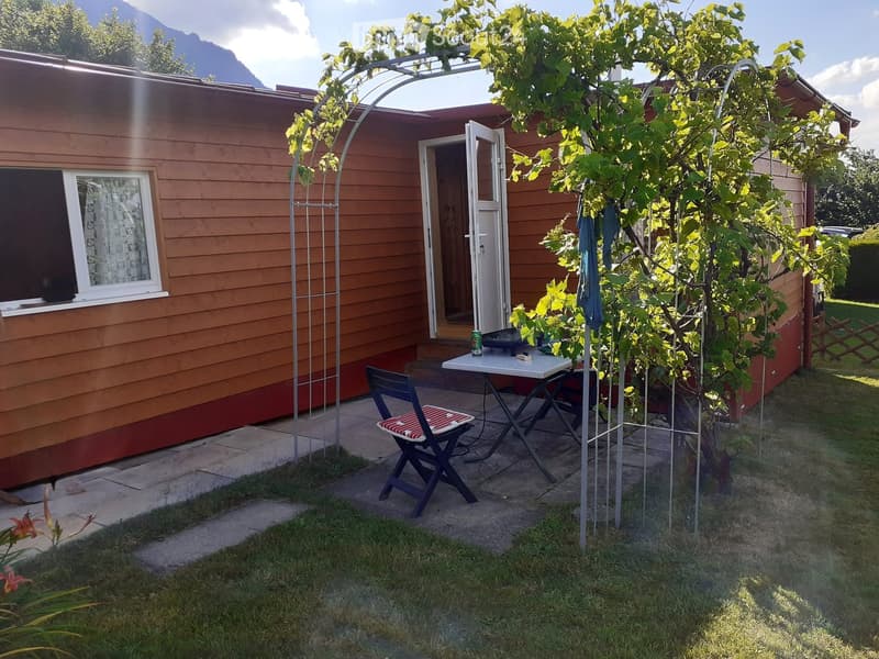 A small outdoor seating area with a table, chair, and umbrella near a wooden house with a window and open door. A tree and metal archway are also visible.