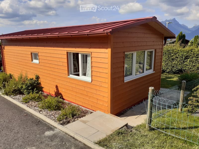Small wooden house, red roof, three windows, small front yard, surrounded by greenery