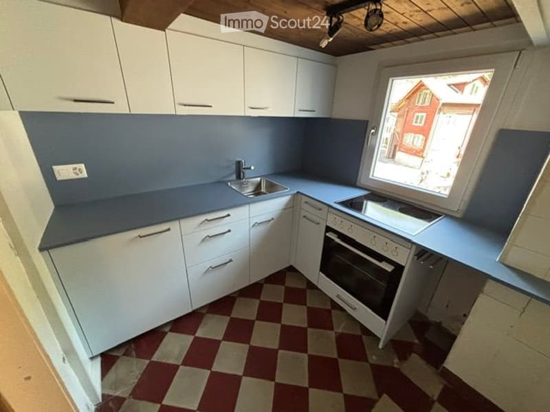 Modern kitchen with white cabinets, blue countertops, and a checkered floor. Features a sink, oven, and a window with a view of a red building.