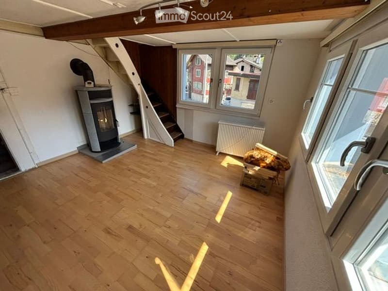 Empty room with wooden flooring, white walls, large windows, and a visible staircase leading to an upper level. A fireplace is present, but no furniture or furnishings are visible.