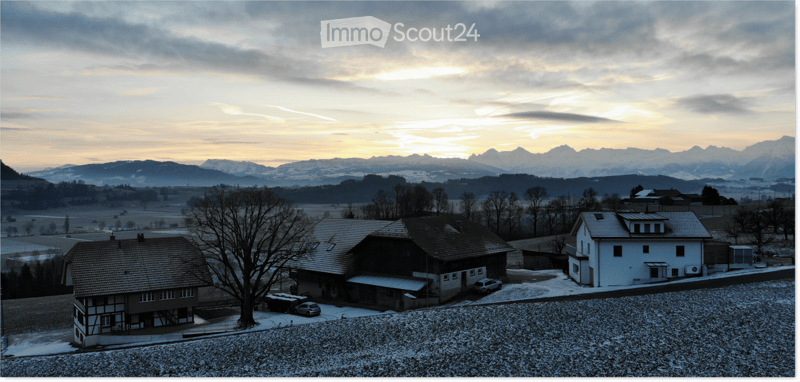 Aerial view of a house with a tiled roof, surrounded by a snow-covered yard and trees, with a scenic mountain backdrop at sunset.