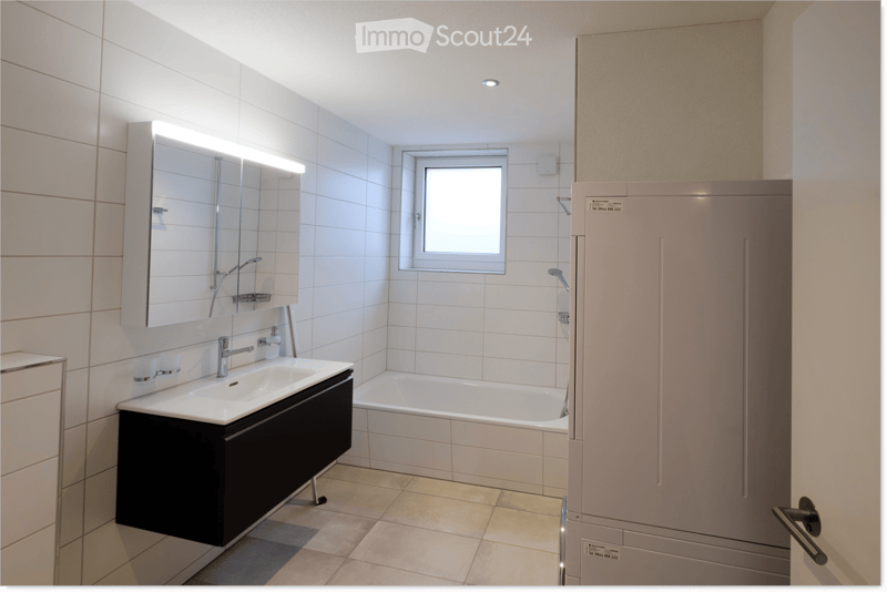 Modern bathroom with white tiles, a black vanity cabinet, a large mirror with built-in lighting, a bathtub, and a window for natural light.