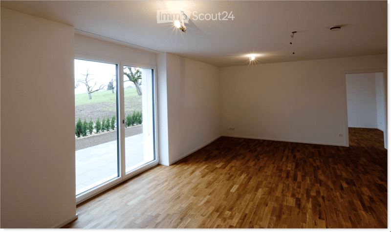 Empty room with wooden flooring, white walls, large sliding glass door leading to a garden, and recessed ceiling lights.
