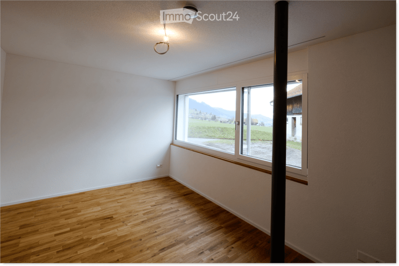 Empty room with wooden flooring, white walls, and large windows offering a scenic view of a green landscape and mountains.