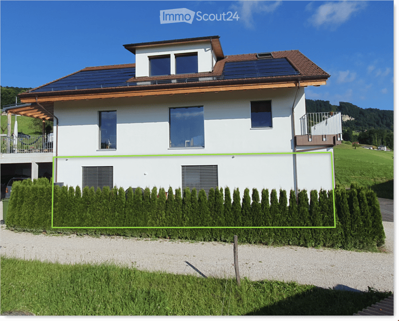 Two-story house with white exterior, large windows, solar panels on the roof, and a garage. The house is surrounded by a neatly trimmed hedge and a gravel driveway.