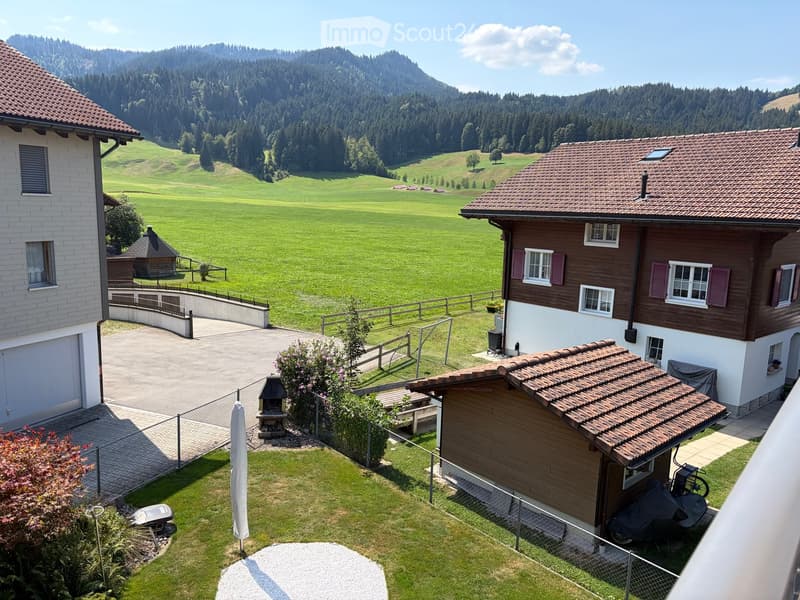 A peaceful garden area with a barbecue, picnic table, and a small wooden shed, surrounded by a fence and lush greenery, with a scenic mountain view in the background.