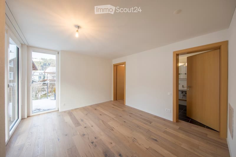 Empty room with light wooden flooring, white walls, and large windows offering a view of the outside. The room features a door leading to another room and a partially visible bathroom with a sink.