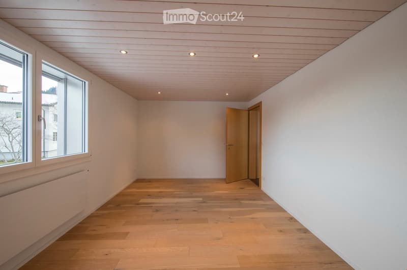 Empty room with light wooden flooring, white walls, and a wooden door leading to another space. Large windows allow natural light to enter.