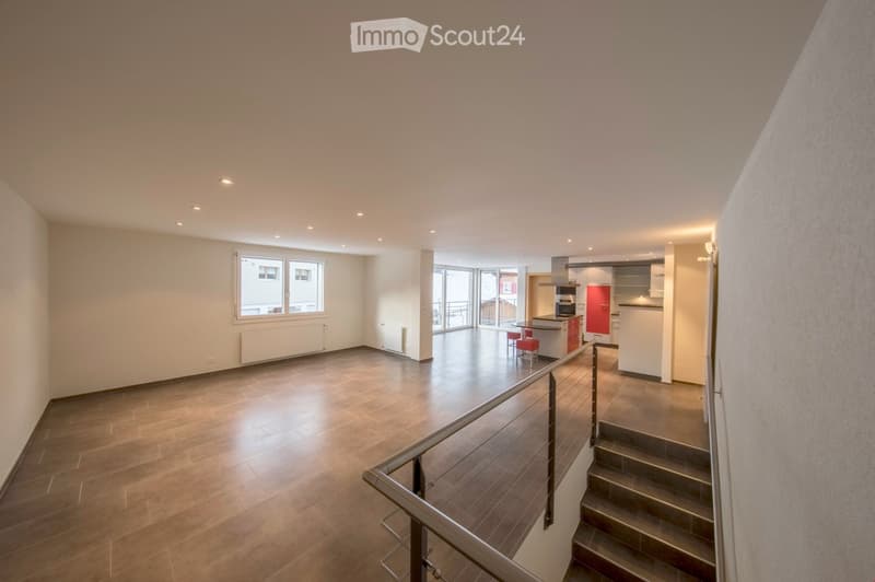 Open-plan living and dining area with wooden flooring, modern kitchen area, two red stools, and a staircase leading to another level.