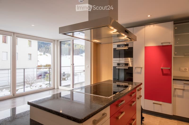 Modern kitchen with white and red cabinets, granite countertop, induction stove, built-in oven, and range hood. Large windows offer a view of the outdoor area.