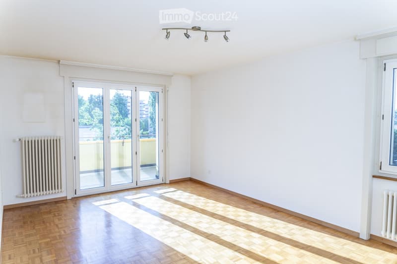 Empty room with wooden flooring, white walls, large glass sliding doors leading to a balcony, radiators on both sides, and a track light fixture on the ceiling.