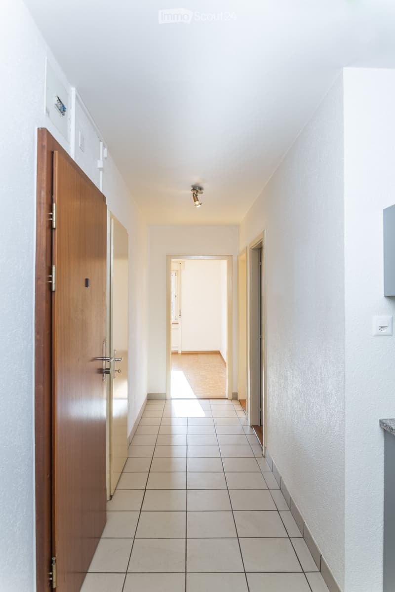 Empty hallway with tiled floor, white walls, and wooden doors on both sides. A ceiling light fixture is visible.