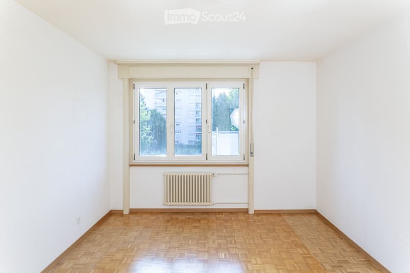Empty room with wooden flooring, white walls, large window with white frame, radiator below the window, and a visible electrical outlet on the left wall.