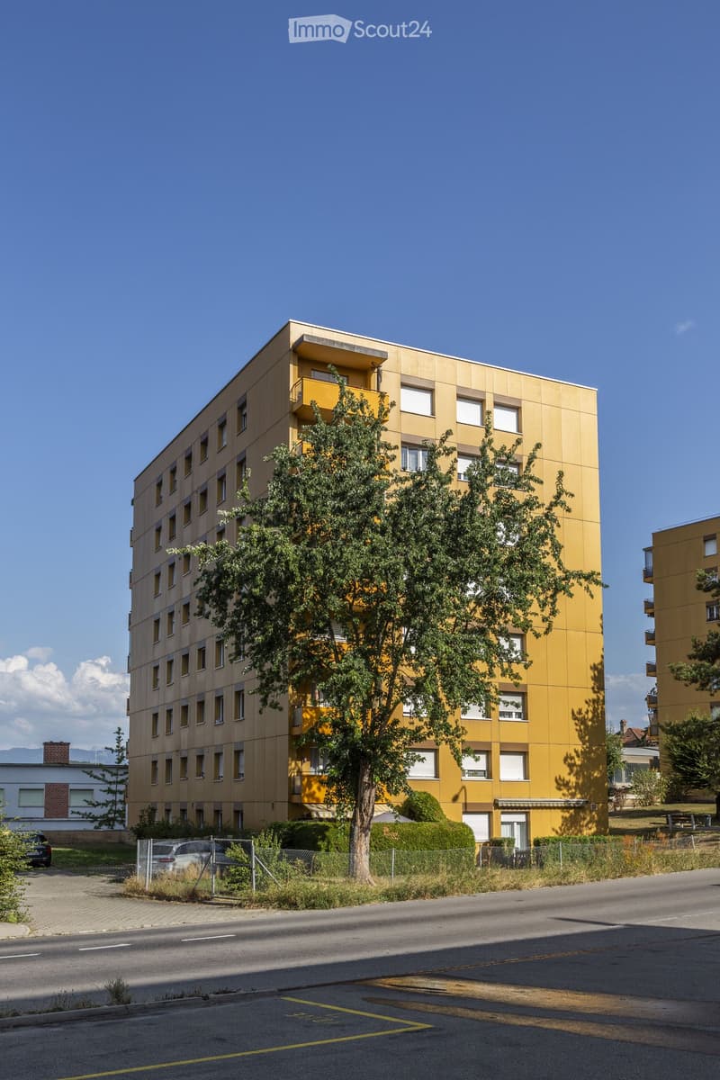 Modern multi-story residential building with yellow facade, multiple windows, and balconies. Surrounded by trees and a parking area.
