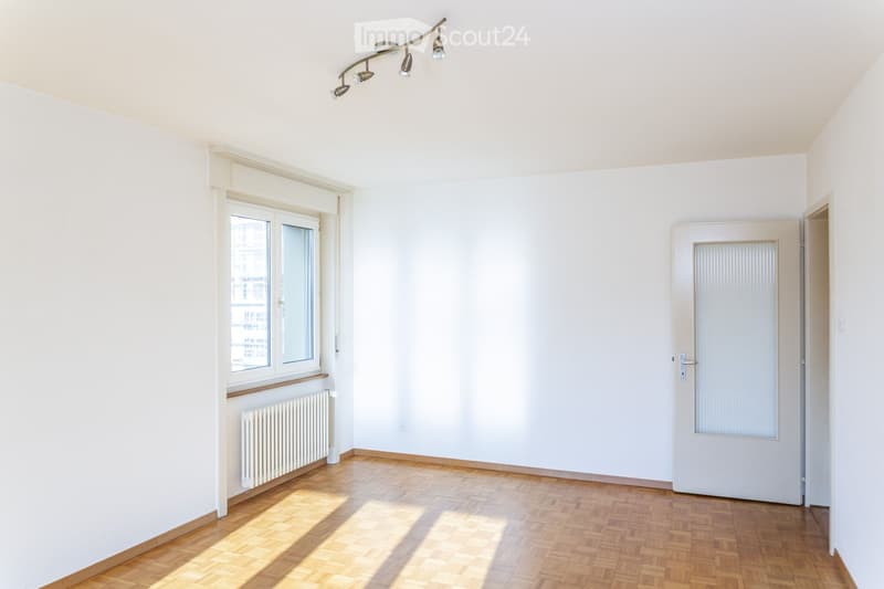 Empty room with white walls, wooden floor, large window, radiator, and a door leading to another space.