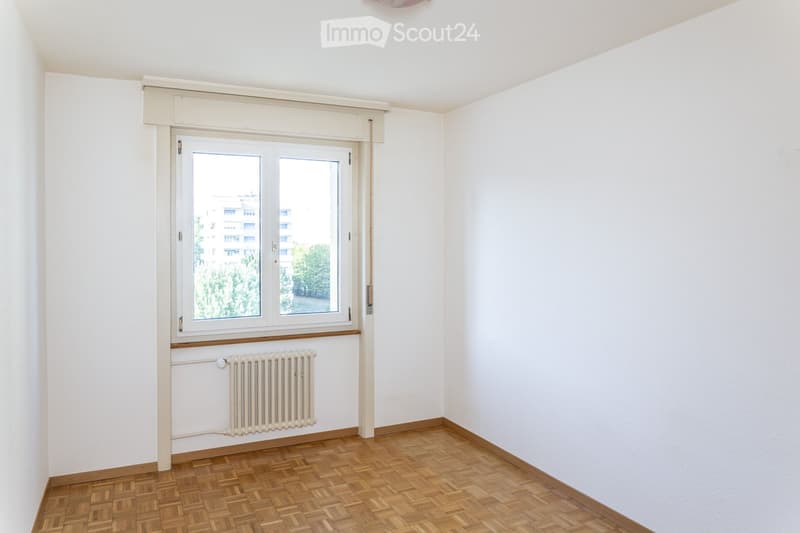 Empty room with white walls, wooden herringbone flooring, and a large window with a radiator below it.